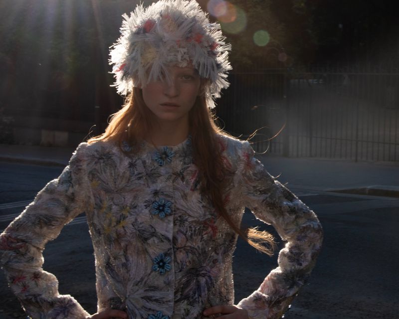 Model wearing a textured patterned jacket and matching feathered hat outdoors during sunset