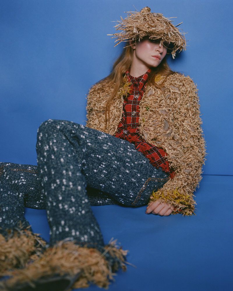 Model seated against blue backdrop wearing a textured straw hat, red plaid shirt, and speckled dark trousers.
