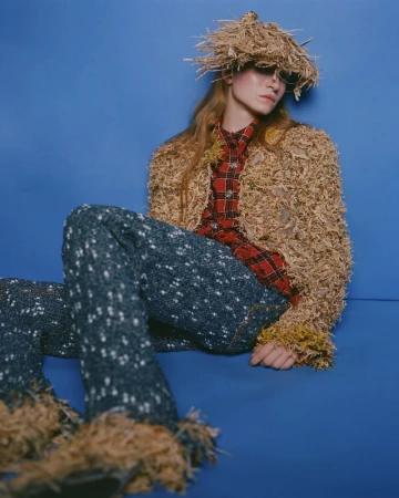 Model seated against blue backdrop wearing a textured straw hat, red plaid shirt, and speckled dark trousers.
