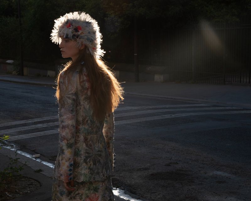 A model standing on a city street wearing a textured patterned outfit and a feathered hat during golden hour.