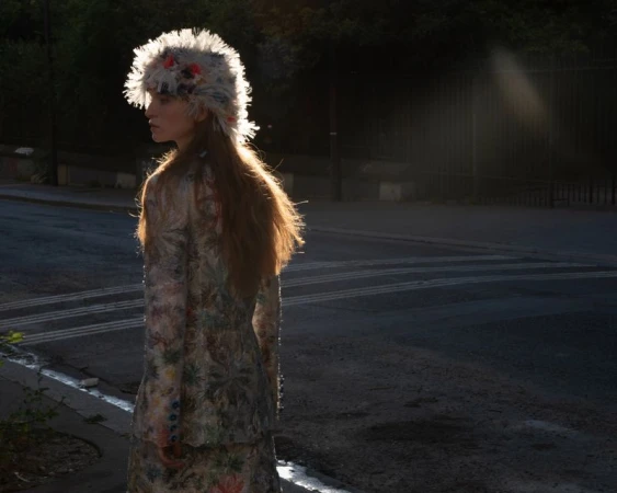 A model standing on a city street wearing a textured patterned outfit and a feathered hat during golden hour.