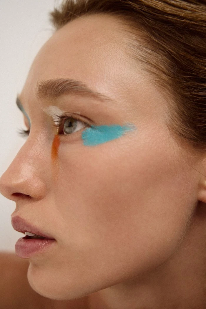 Close-up portrait of a woman with vibrant blue and orange makeup strokes on her face against a plain background