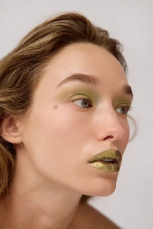 Close-up beauty portrait of a woman wearing metallic green lipstick and olive eyeshadow against a neutral white background