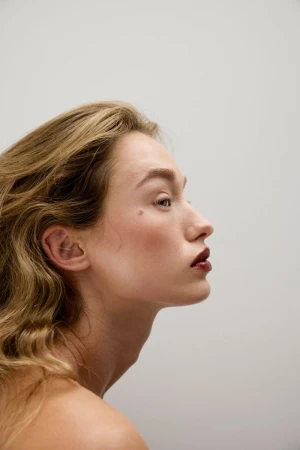 Profile portrait of a model with blonde hair and dark glossy lipstick against a neutral light grey background.