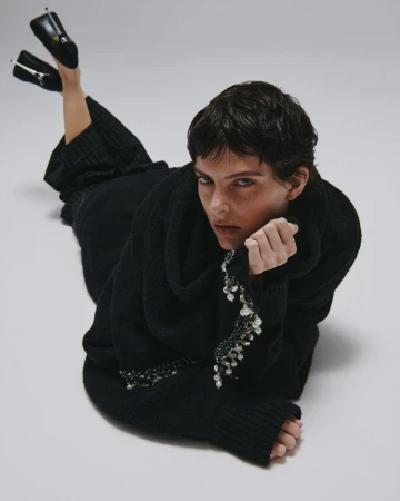 Model in black sweater with pearl embellishments lying on a white studio backdrop, wearing dark heels