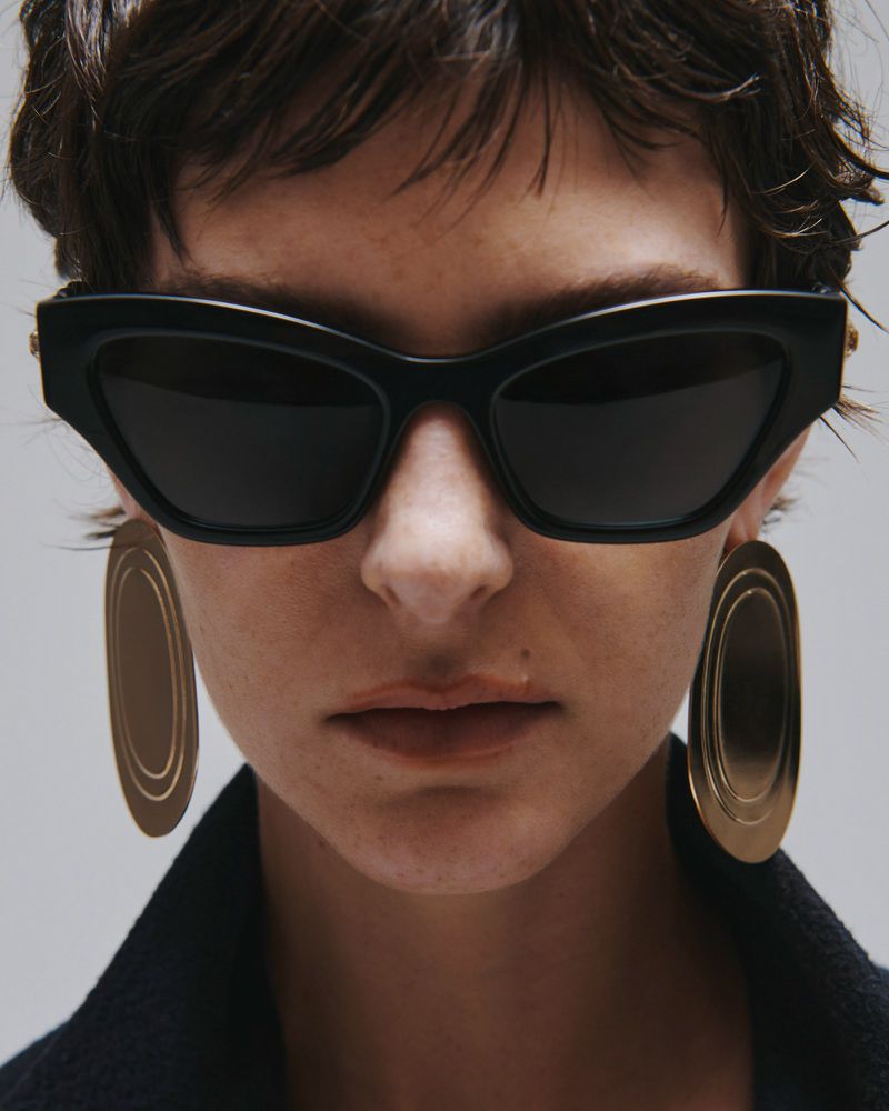 Close-up portrait of a model wearing dark sunglasses and large metallic earrings against a neutral grey background.