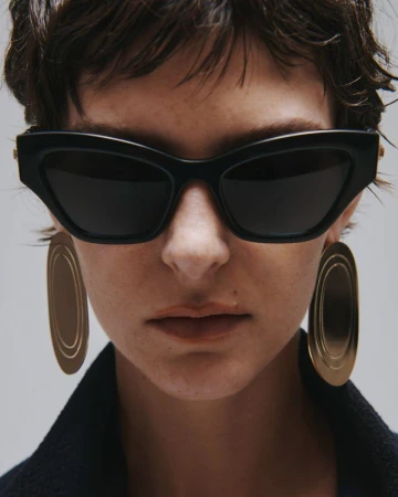 Close-up portrait of a model wearing dark sunglasses and large metallic earrings against a neutral grey background.