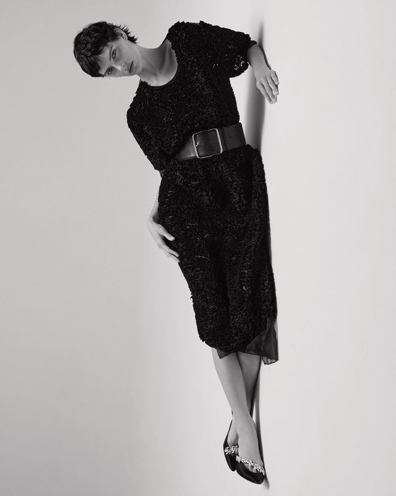 Model with short dark hair in black textured dress and wide belt posing against white seamless studio backdrop