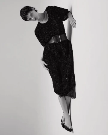 Model with short dark hair in black textured dress and wide belt posing against white seamless studio backdrop