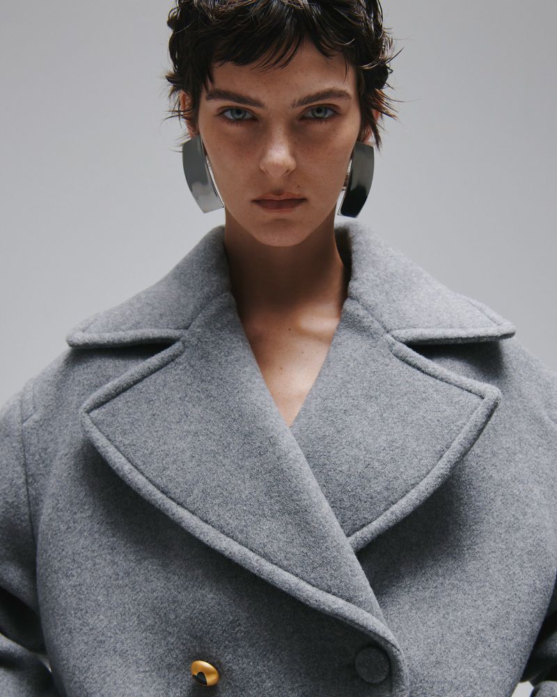 Model with dark short hair wearing a grey wool coat and large metallic earrings against a grey background.