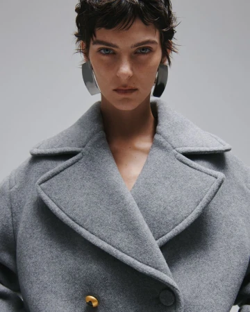 Model with dark short hair wearing a grey wool coat and large metallic earrings against a grey background.