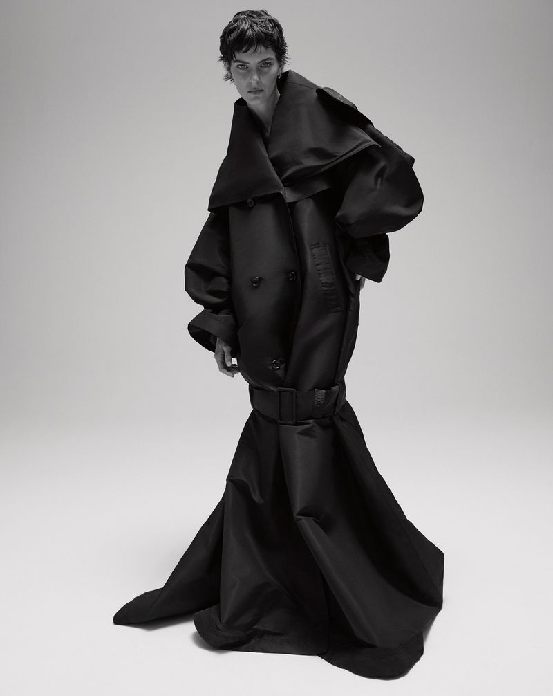 Model with dark short hair wearing an oversized black sculptural belted coat against a plain studio background.