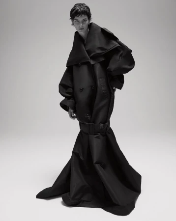 Model with dark short hair wearing an oversized black sculptural belted coat against a plain studio background.