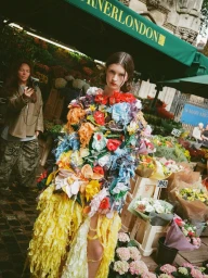 Model wearing a vibrant floral-encrusted garment standing in an outdoor flower market stall