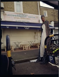 Model standing outdoors in front of a London storefront wearing a patterned polka-dot dress and matching headwrap.