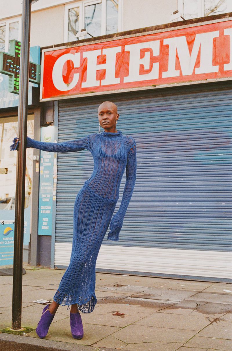 Model wearing a blue knit dress and purple boots standing before a storefront with a chemist sign