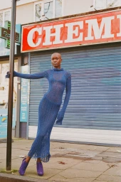 Model wearing a blue knit dress and purple boots standing before a storefront with a chemist sign