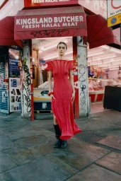 Model in a red dress posing on a city pavement in front of a red storefront butcher shop