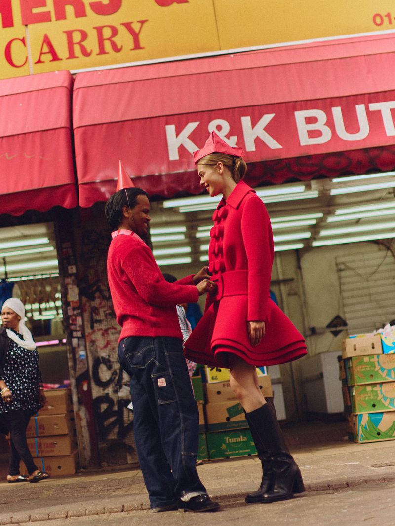 Model in a structured red coat and matching hat standing outside a storefront with a person adjusting her belt