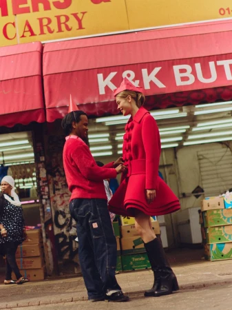 Model in a structured red coat and matching hat standing outside a storefront with a person adjusting her belt
