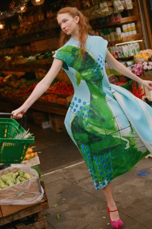 Model in a structured blue and green patterned dress holding a green shopping basket inside a market