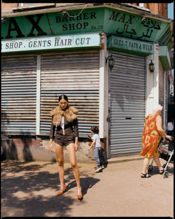 Model standing in front of a barbershop storefront wearing a brown jacket with fur collar, shorts, and heels.