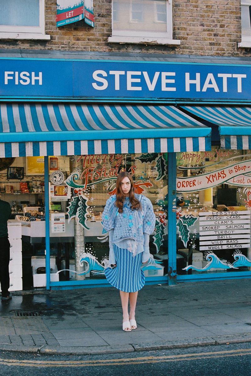 Model standing in front of a blue storefront awning wearing patterned blue textured clothing and white shoes.