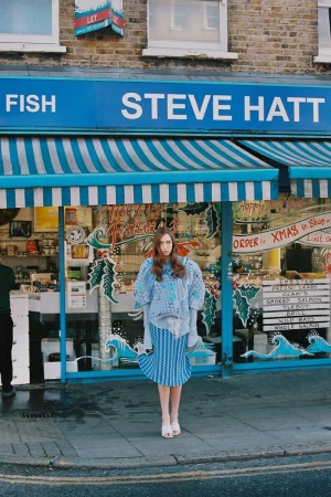 Model standing in front of a blue storefront awning wearing patterned blue textured clothing and white shoes.