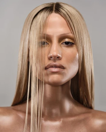 Close up portrait of model Gabbriette with straight blonde hair falling over her face against a neutral grey background.