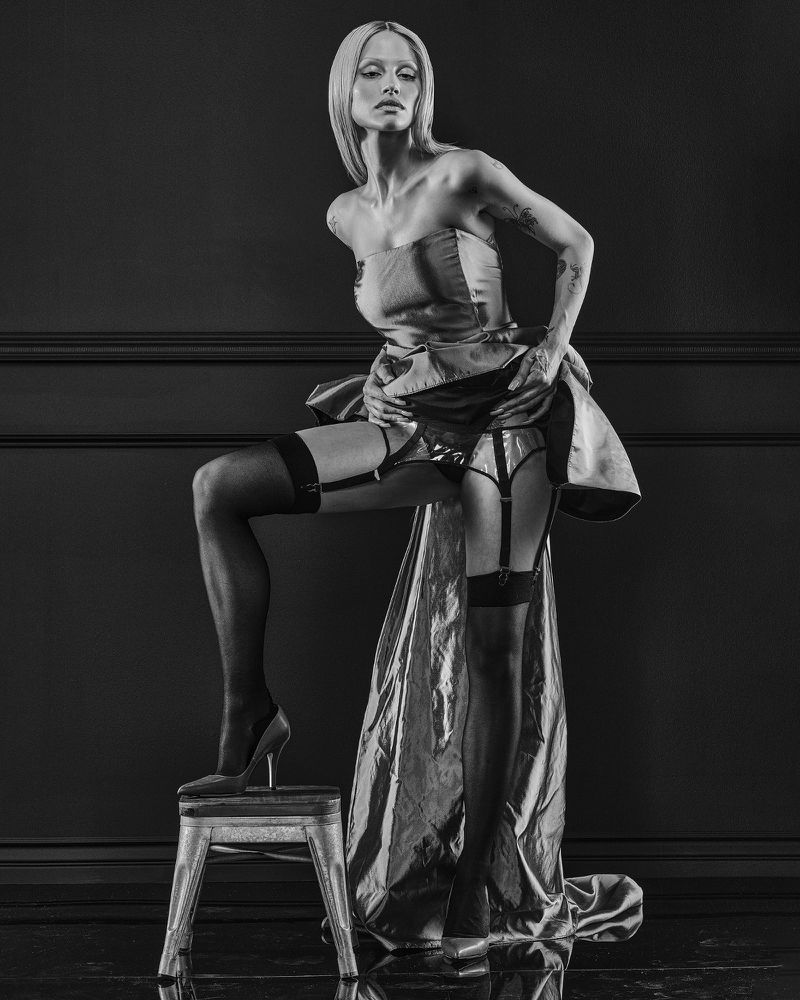 Model Gabbriette posing in lingerie and a structured dress standing with one foot on a metal stool against a dark background
