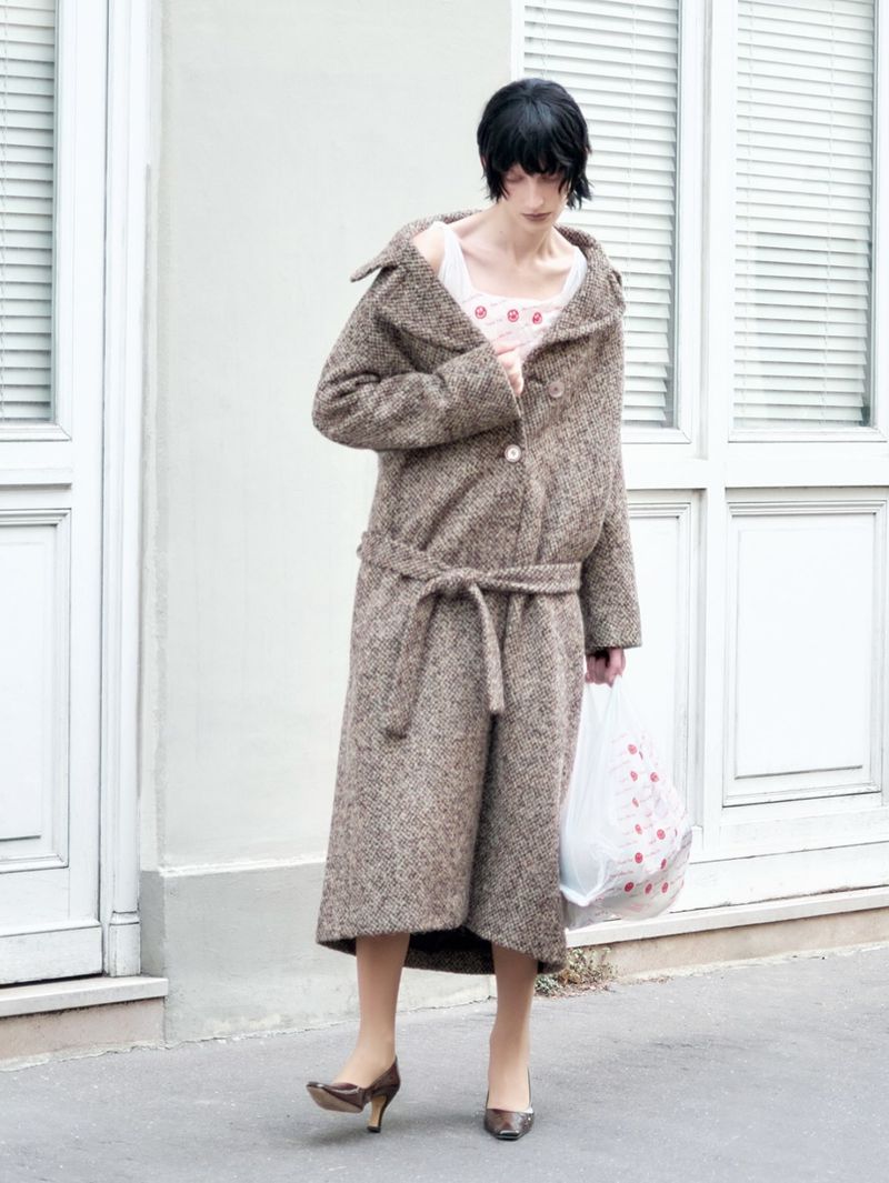 Model in brown tweed belted coat and heels holding plastic bag standing outdoors against a pale building exterior