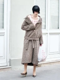 Model in brown tweed belted coat and heels holding plastic bag standing outdoors against a pale building exterior