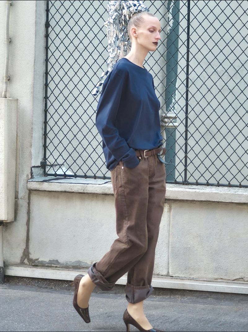 Model wearing a blue top and brown trousers posing against a chain-link fence outdoors for D Repubblica editorial.