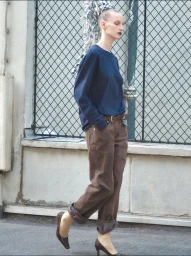 Model wearing a blue top and brown trousers posing against a chain-link fence outdoors for D Repubblica editorial.