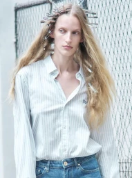 Model Kira Alferink with metal hair clips in long blonde hair wearing a striped shirt and denim jeans against a fence.