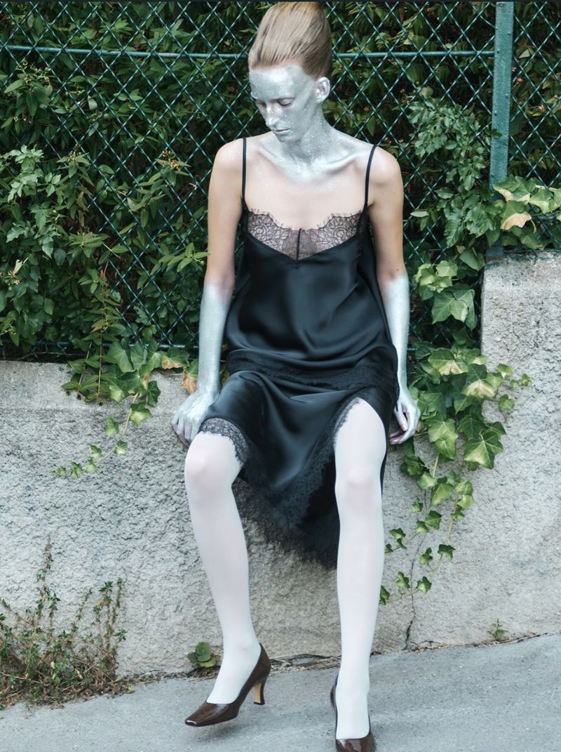 Model with silver body paint wearing a black slip dress and white tights sitting against a low concrete wall with a chain-link fence.