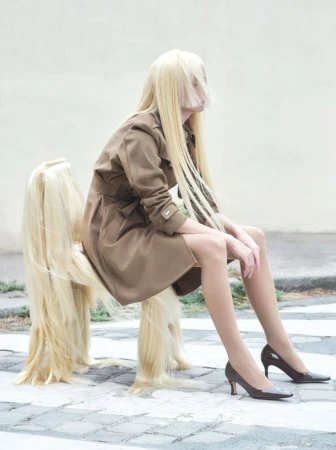 Blonde model wearing a brown belted coat and dark heels sitting on a hair-covered chair outdoors.