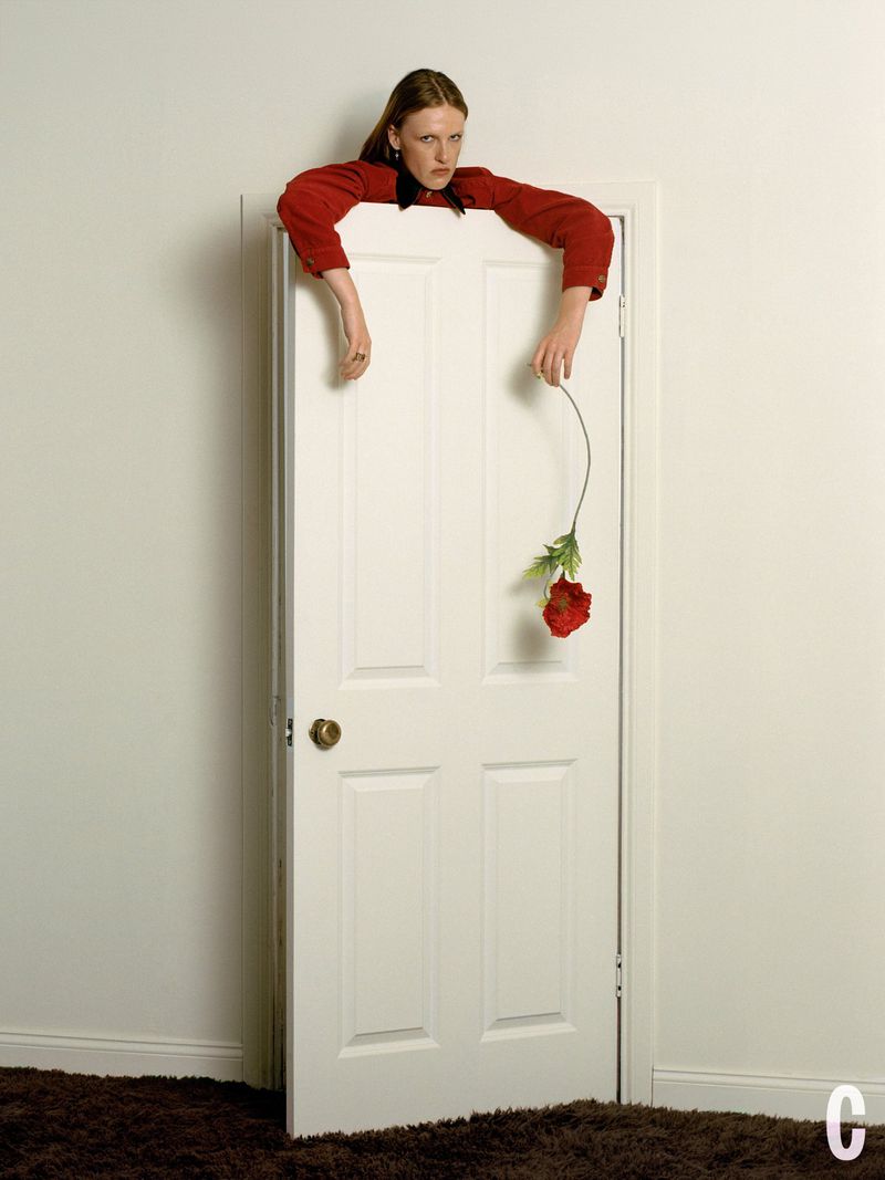 Woman in a red jacket leaning over the top of a white closed door holding a flower for Cosmopolitan US