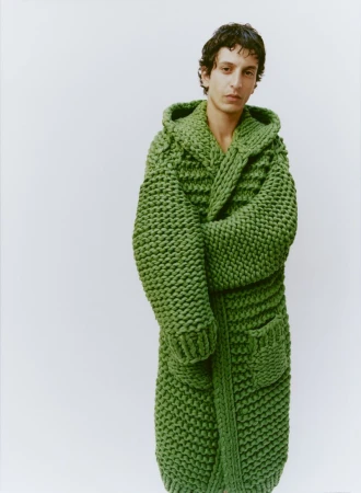 Model wearing a thick chunky-knit green hooded cardigan against a plain white studio background