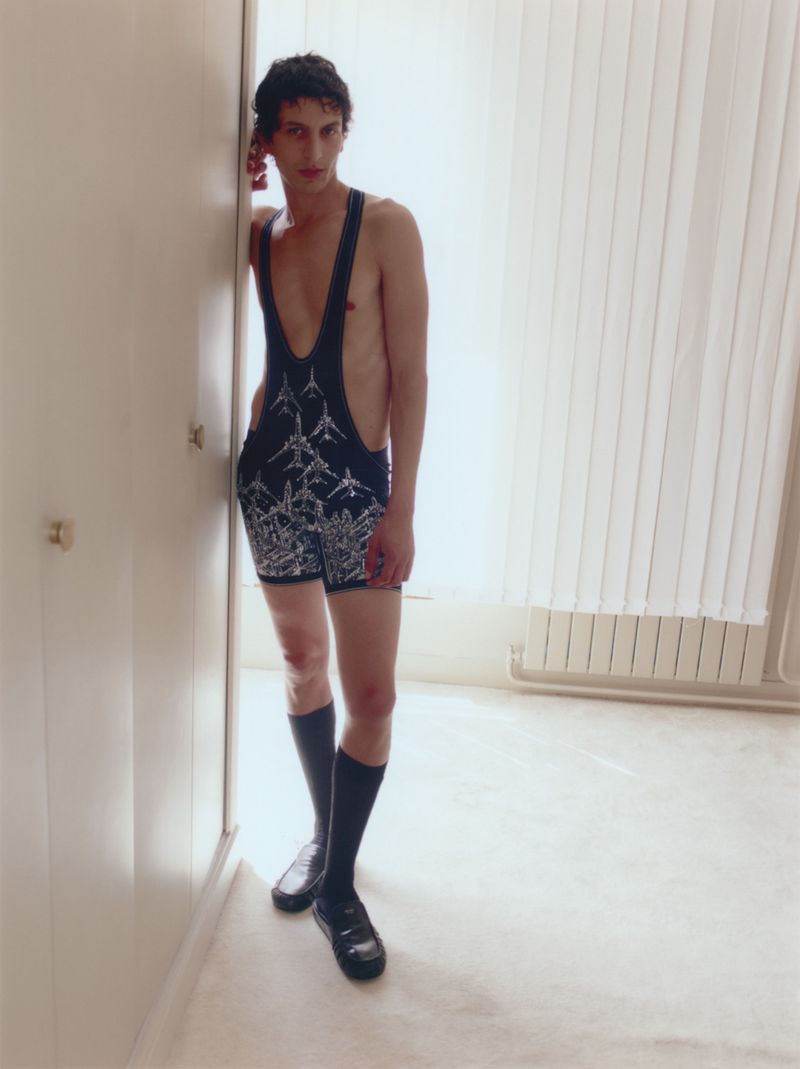 Model standing indoors wearing a printed dark romper with high socks and loafers against a pale interior background.