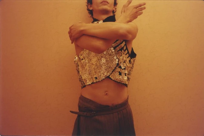 Man wearing a sequined crop top and belted trousers standing against a warm wall