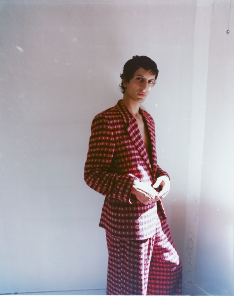 Man wearing a red patterned suit standing against a plain white wall in soft natural light.