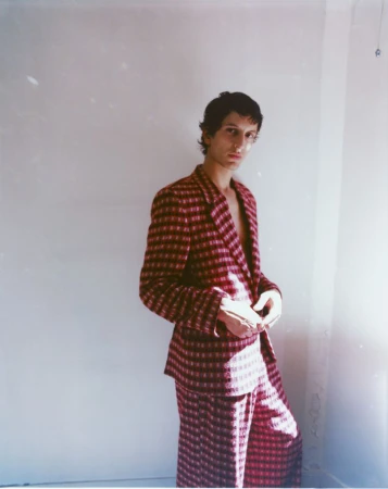 Man wearing a red patterned suit standing against a plain white wall in soft natural light.