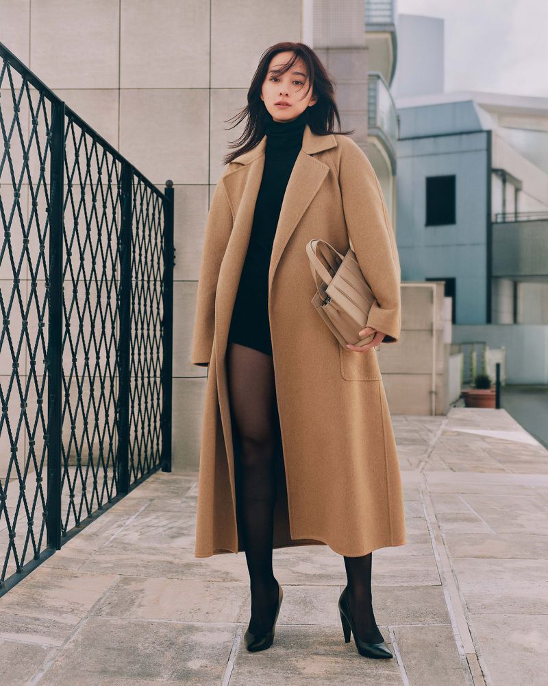 Woman wearing a long camel coat and black heels holding a handbag outdoors on a paved street.