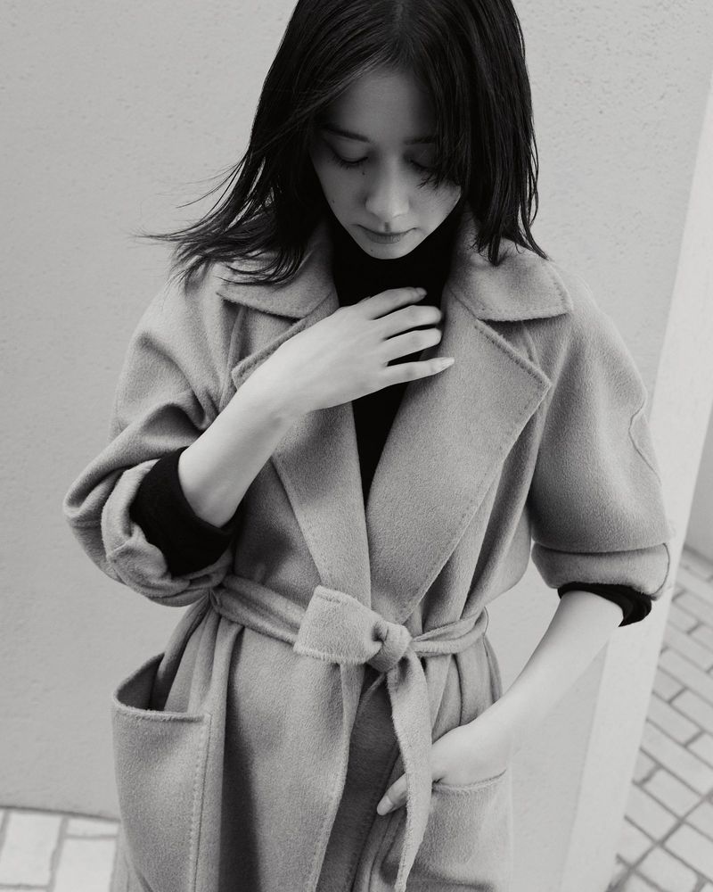 Monochrome portrait of a woman wearing a belted wool coat and dark turtleneck sweater against an outdoor wall.