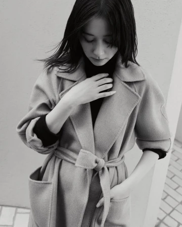 Monochrome portrait of a woman wearing a belted wool coat and dark turtleneck sweater against an outdoor wall.