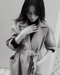 Monochrome portrait of a woman wearing a belted wool coat and dark turtleneck sweater against an outdoor wall.