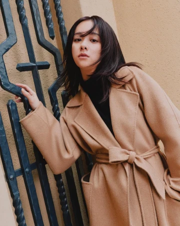 Woman in a camel belted wool coat leaning against a dark wrought iron gate and textured wall