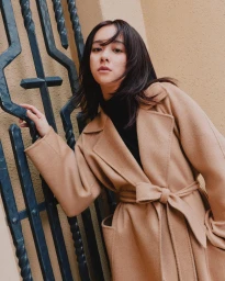 Woman in a camel belted wool coat leaning against a dark wrought iron gate and textured wall