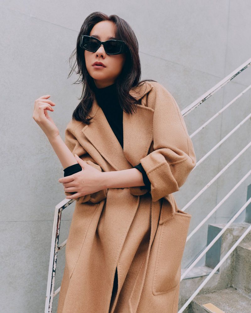 Model wearing dark sunglasses and a long camel wool coat posing on outdoor stairs with a white railing for Max Mara.